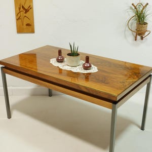 Peut inclure: Table basse rectangulaire en bois avec une finition brillante, soutenue par des pieds en métal. La table présente un arrangement décoratif d'une petite plante en pot et de deux vases marron sur une nappe en dentelle. Une tenture murale est visible en arrière-plan.