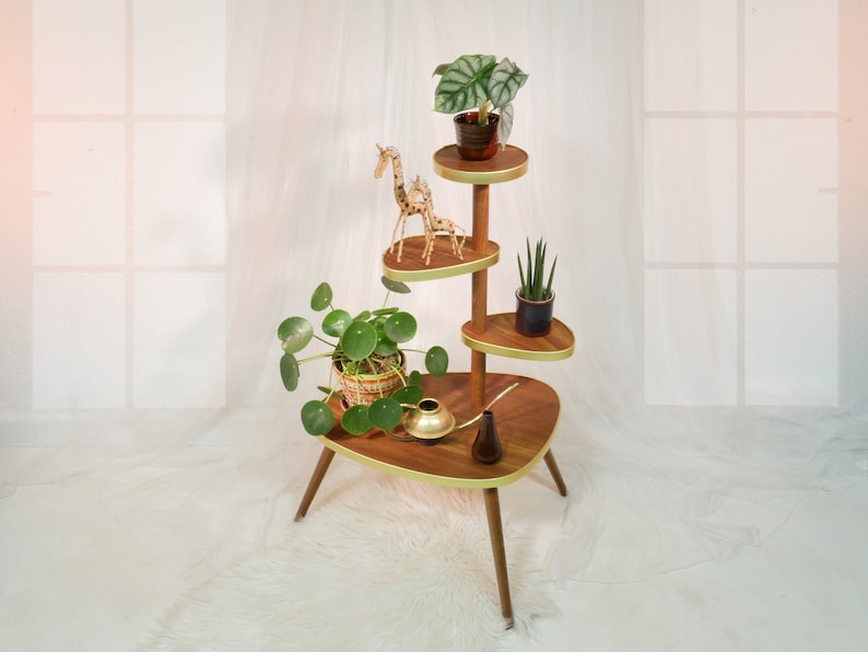 May include: A mid-century modern plant stand with four tiers, featuring a gold trim and a wooden base. The stand is decorated with a wooden giraffe figurine and potted plants.
