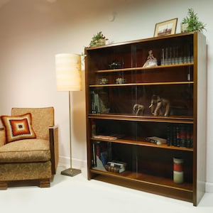 Pode incluir: Uma estante moderna de meados do século com portas de correr de vidro, cheia de livros e objetos decorativos. Uma poltrona estampada com uma almofada quadrada e um candeeiro de pé estão ao lado da estante. A estante é de madeira escura.