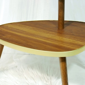 May include: A mid-century modern side table with a wood top and gold trim. The table has three legs and a rounded, oval-shaped top.