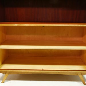 May include: A mid-century modern wooden cabinet with two doors open, revealing three shelves. The cabinet has a dark brown top and sides, with a lighter wood interior and shelves. The cabinet is supported by angled legs.