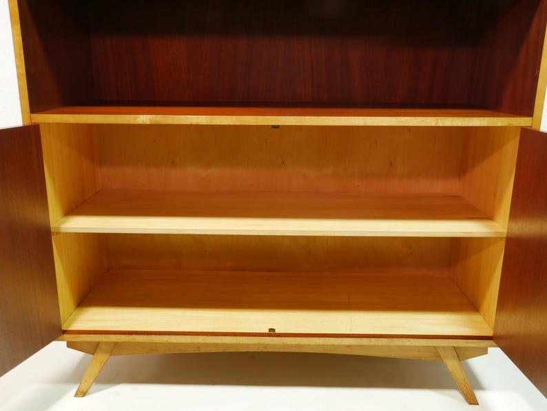 May include: A mid-century modern wooden cabinet with two doors open, revealing three shelves. The cabinet has a dark brown top and sides, with a lighter wood interior and shelves. The cabinet is supported by angled legs.