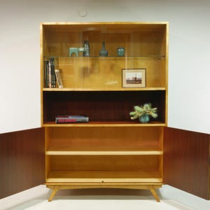 May include: A mid-century modern wooden cabinet with open doors. The cabinet has a light wood finish with a dark wood interior. It features shelves, a glass display area, and decorative items.