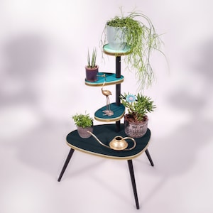 Op de afbeelding: Een turquoise plantenstandaard met vier niveaus en zwarte poten. De standaard heeft gouden accenten en een messing gieter. De bovenste laag heeft een lichtblauwe pot met een groene plant. De tweede laag heeft een paarse pot met een groene plant. De derde laag heeft een gouden flamingo-beeldje. De onderste laag heeft een bruine pot met een groene plant.