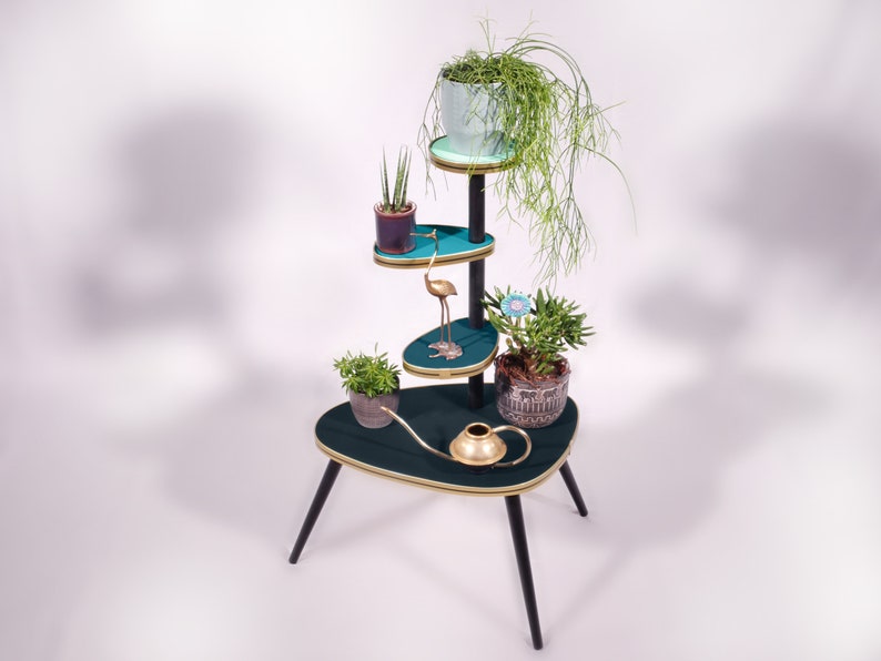 Op de afbeelding: Een turquoise plantenstandaard met vier niveaus en zwarte poten. De standaard heeft gouden accenten en een messing gieter. De bovenste laag heeft een lichtblauwe pot met een groene plant. De tweede laag heeft een paarse pot met een groene plant. De derde laag heeft een gouden flamingo-beeldje. De onderste laag heeft een bruine pot met een groene plant.