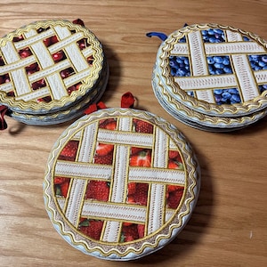 May include: Three round fabric pot holders with a pie crust design. One pot holder has a cherry pie design, one has a blueberry pie design, and one has a strawberry pie design.