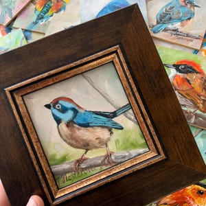 May include: A framed painting of a blue and brown bird perched on a branch. The bird has a blue head and wings, with a brown body. The painting is in a dark brown wooden frame with a gold inner border. Other bird paintings are in the background.