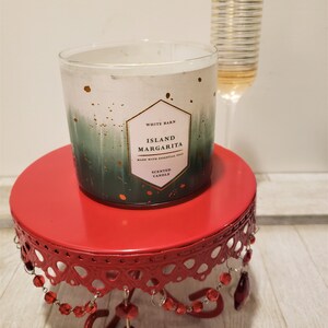 May include: A white candle with a green and gold ombre design. The candle is labeled "White Barn Island Margarita Scented Candle" and is sitting on a red metal cake stand with red beads.