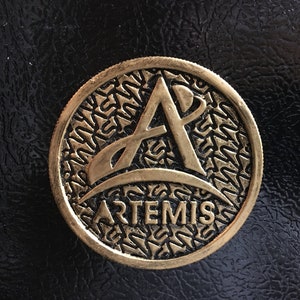 May include: A bronze-colored Artemis coin with a textured border and the Artemis logo in the center. The coin is set against a black, textured background. The word "ARTEMIS" is inscribed below the logo.