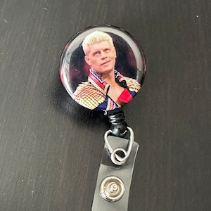 Könnte beinhalten: Ein schwarz-weißer Knopf mit einem Foto eines Mannes mit blonden Haaren, der ein goldenes und rotes Kostüm trägt. Der Knopf ist an einem durchsichtigen, einziehbaren Ausweishalter aus Kunststoff mit einem silbernen Clip und Knopf befestigt.