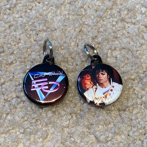 May include: Two round keychains with metal rings. One keychain features a black and red logo with the text "CAPTAIN EO" in white. The other keychain features a color photo of Michael Jackson with a white cat on his shoulder.