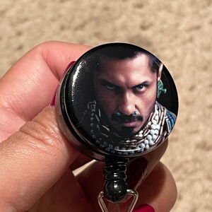 May include: A black retractable badge reel with a round, black button featuring a color photograph of a man with dark hair and a beard. The man is wearing a dark shirt and a silver necklace.