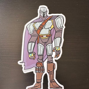 Puede incluir: Una pegatina con una ilustración de dibujos animados de un guerrero mandaloriano con una capa morada y armadura plateada. El personaje sostiene un rifle bláster y tiene un símbolo azul y amarillo en su brazo. La pegatina tiene el texto "@MIGUELWILSON" en la parte inferior.
