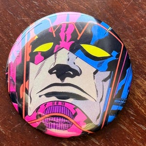 May include: A circular pinback button featuring a stylized graphic illustration of a comic book character with a pale face, bright yellow eyes, and a purple chin piece. The design incorporates vibrant neon pink and blue accents against a dark background.