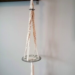 Pode incluir: Suporte de planta em macramé creme com base de engrenagem de bicicleta de metal prateado. O suporte é suspenso por um suporte de metal em uma parede branca. Uma borla pende da parte inferior da engrenagem.