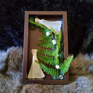 May include: A framed shadow box featuring two white ceramic dress forms, green fern fronds, and small white flower accents. The brown frame provides a rustic contrast to the delicate arrangement, creating a unique decorative piece.