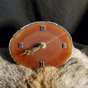 May include: A round clock made from a polished, brown agate slice. The clock face features Roman numerals and gold-colored hands. The clock is a decorative item.