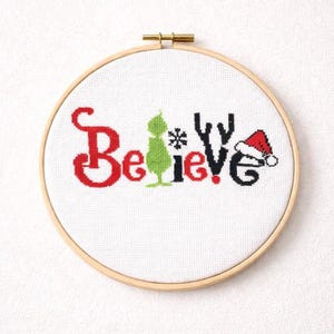 Op de afbeelding: Een borduurring met kruissteek met het woord "Believe" in rood en zwart, met een groene Grinch-achtige figuur en een kerstmuts. Het ontwerp staat op witte stof, gezet in een houten ring met een messing sluiting. Een feestelijke vakantiedecoratie.