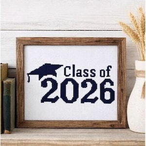 May include: A framed cross-stitch artwork with the text "Class of 2026" in navy blue, featuring a graduation cap. The artwork is in a brown wooden frame, displayed on a wooden shelf with books and a vase.