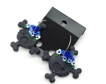 May include: A pair of black skull earrings with blue and white flower accents. The earrings are made of polymer clay and have a hoop design.