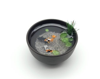 Puede incluir: Un cuenco de cerámica negro en miniatura lleno de resina, que representa un pequeño estanque con dos peces koi naranja y negro, plantas verdes y grava gris.