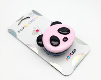 May include: Pink phone grip with a panda face design. The grip has a black outline and black eyes. The packaging says "PopGrip" and "Support-Poigneé De Téléphone Grip".