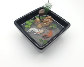 Puede incluir: Un estanque en miniatura de resina con dos peces dorados, plantas verdes y pequeñas piedras en un plato cuadrado negro.