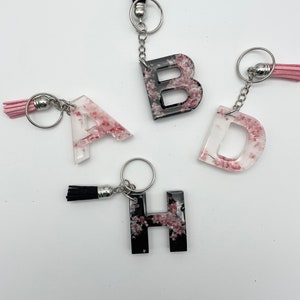 Puede incluir: Cuatro llaveros de resina con forma de letras con purpurina negra y rosa. Las letras son A, B, D y H. Cada llavero tiene un anillo de metal plateado y un borla negra.