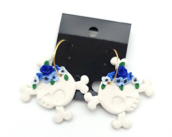May include: A pair of white skull earrings with blue and white flower accents. The earrings are made of polymer clay and have gold hoop closures.