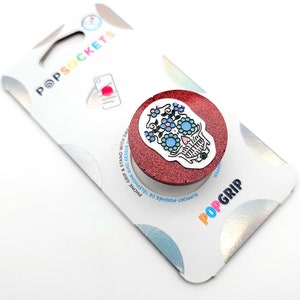 Puede incluir: Un agarre para teléfono de glitter rojo con un diseño de calavera de azúcar blanca. La calavera tiene flores azules y rosas y un corazón verde.