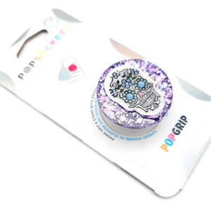 Puede incluir: Agarradera para teléfono de purpurina morada con un diseño de calavera de azúcar blanca. La calavera tiene flores azules y un corazón rosa en la frente. El texto "POPGRIP" está impreso en el empaque.