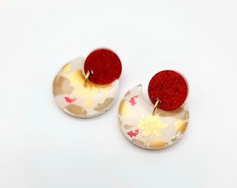 May include: A pair of earrings with a crescent moon shape and a red circle. The crescent moon is white with a gold and red floral pattern. The red circle is a solid color.
