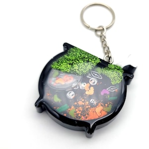 May include: Black cauldron keychain with green glitter and Halloween-themed charms inside. The cauldron is filled with orange, green, and black glitter.