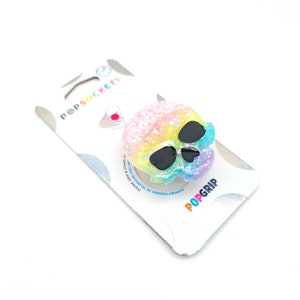 May include: A rainbow glitter skull phone grip with black sunglasses. The grip is attached to a white card with the text "POPSOCKET" and "POPGRIP".