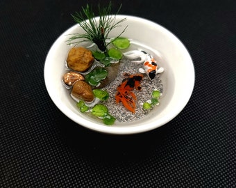 Puede incluir: Un estanque de resina en miniatura con dos peces dorados naranja y blancos, plantas verdes y pequeñas piedras marrones en un cuenco blanco.