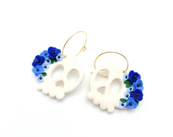 May include: A pair of white ceramic skull earrings with blue and light blue flower accents. The earrings are on a white background.