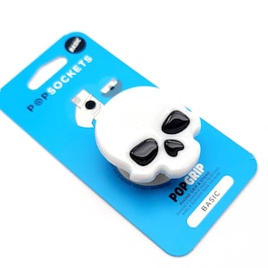 May include: White skull-shaped PopSockets phone grip with black eye sockets, attached to a blue card. The card has the PopSockets logo and the words "PHONE GRIP & STAND" and "BASIC".