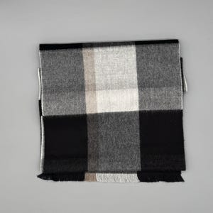 May include: A rectangular scarf featuring a large-scale plaid pattern in shades of black, white, and gray. The fabric has a visible woven texture and the edges are finished with short, straight fringe detailing along the bottom hemline.