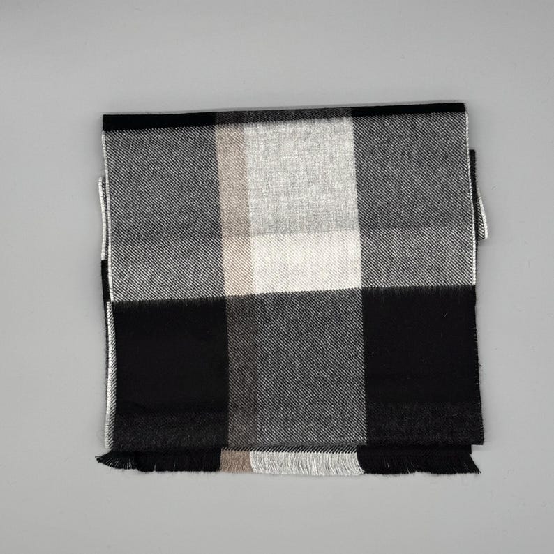 May include: A rectangular scarf featuring a large-scale plaid pattern in shades of black, white, and gray. The fabric has a visible woven texture and the edges are finished with short, straight fringe detailing along the bottom hemline.