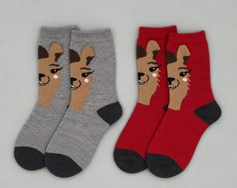 May include: Two pairs of crew socks, one pair in heather gray and one pair in deep red. Each sock features a tan llama face graphic with dark brown ears, black eyelashes, and a small pink cheek detail. The heels and toes are dark charcoal gray.
