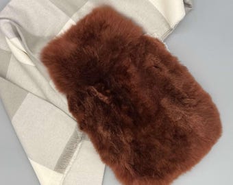 May include: A rectangular hot water bottle cover made of thick, rust-colored faux fur. The cover is positioned over a folded, light gray and white checkered scarf featuring a soft, woven texture and frayed edges along the bottom hem.