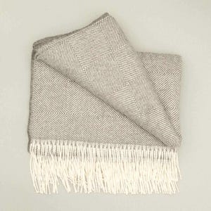 Può includere: Una coperta piegata grigia e bianca con un motivo a rombi. La coperta ha una frangia di nappe bianche lungo un bordo. La coperta è realizzata in un materiale morbido e tessuto.