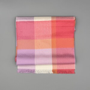 May include: A rectangular scarf featuring a large-scale plaid pattern in shades of pink, orange, lavender, and cream. The fabric has a visible diagonal weave texture and is finished with short, straight fringe along the bottom edge.