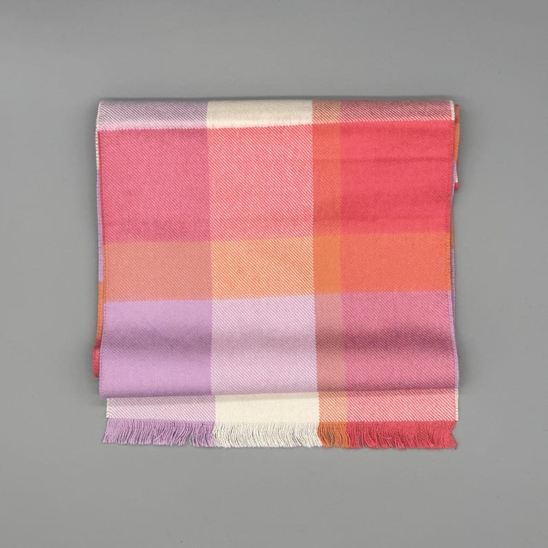 May include: A rectangular scarf featuring a large-scale plaid pattern in shades of pink, orange, lavender, and cream. The fabric has a visible diagonal weave texture and is finished with short, straight fringe along the bottom edge.