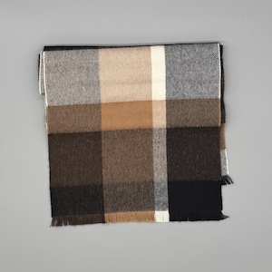 May include: A rectangular scarf featuring a large-scale plaid pattern in shades of black, dark brown, tan, cream, and grey. The fabric has a visible diagonal weave texture and is finished with short, straight fringe along the bottom edge.