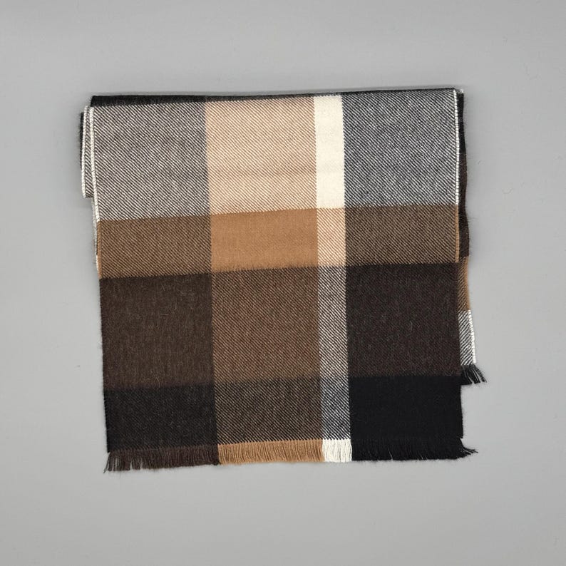 May include: A rectangular scarf featuring a large-scale plaid pattern in shades of black, dark brown, tan, cream, and grey. The fabric has a visible diagonal weave texture and is finished with short, straight fringe along the bottom edge.