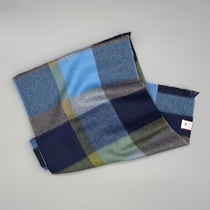 May include: A rectangular scarf featuring a large-scale plaid pattern in shades of navy blue, light blue, olive green, and grey. The fabric has a soft texture with fringed edges on the ends and a small white rectangular brand label attached.