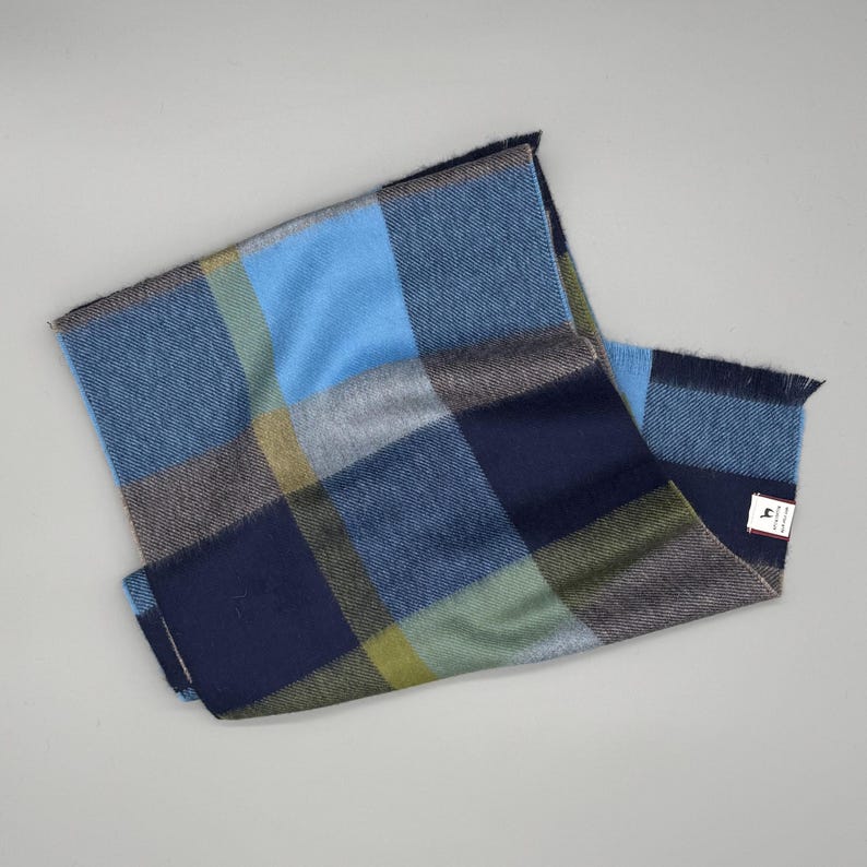 May include: A rectangular scarf featuring a large-scale plaid pattern in shades of navy blue, light blue, olive green, and grey. The fabric has a soft texture with fringed edges on the ends and a small white rectangular brand label attached.