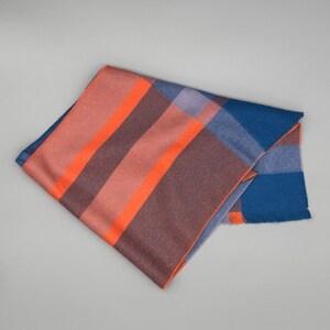 May include: A folded rectangular scarf featuring a plaid pattern with bold stripes in bright orange, muted mauve, deep blue, and soft lavender. The fabric has a smooth texture and a straight, finished edge with a subtle fringe detail on one side.