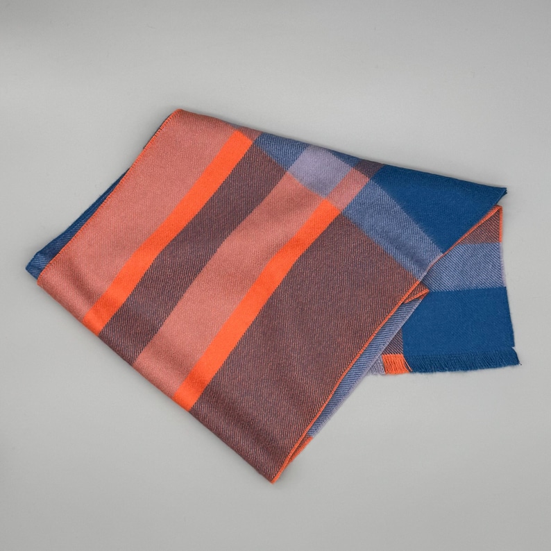 May include: A folded rectangular scarf featuring a plaid pattern with bold stripes in bright orange, muted mauve, deep blue, and soft lavender. The fabric has a smooth texture and a straight, finished edge with a subtle fringe detail on one side.
